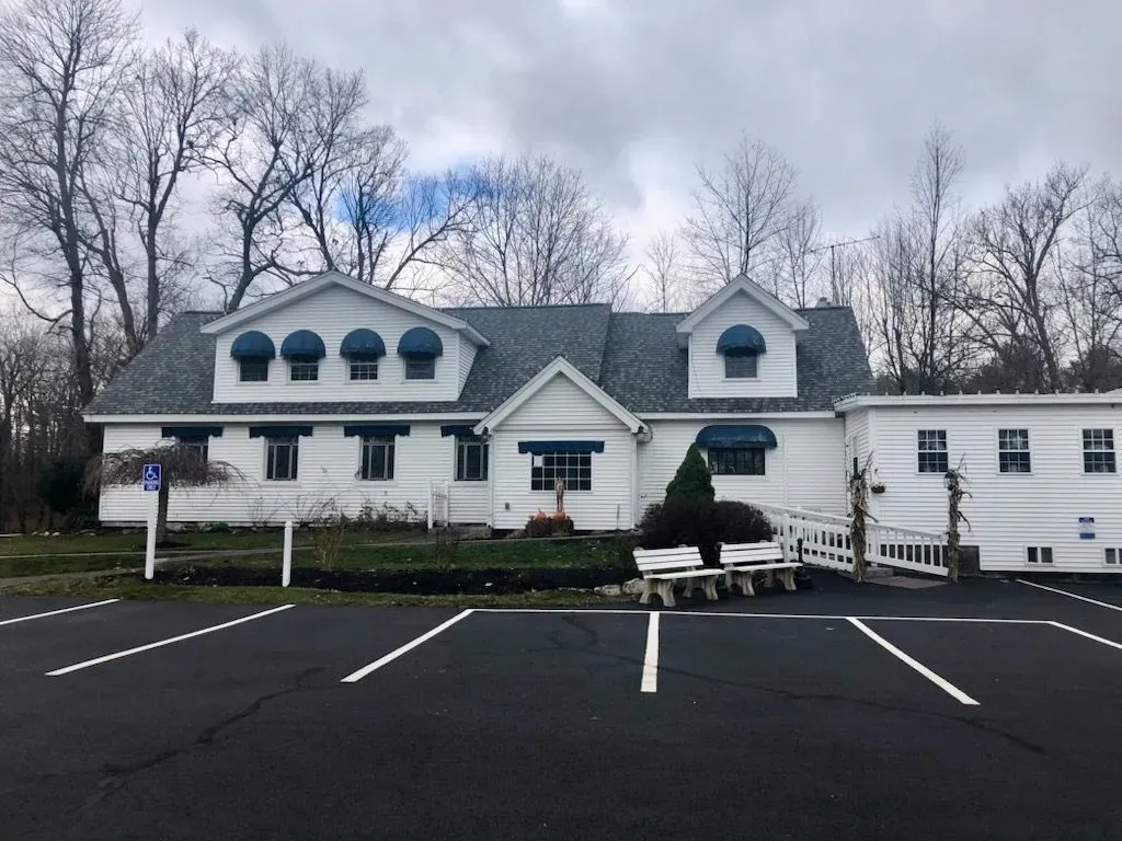 White building with green roof, dormers, and parked car in front of an empty lot under cloudy trees