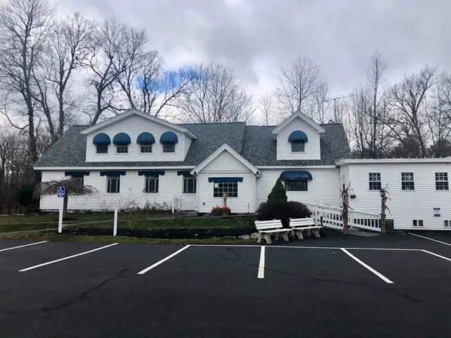 White building with green roof, dormers, and parked car in front of an empty lot under cloudy trees