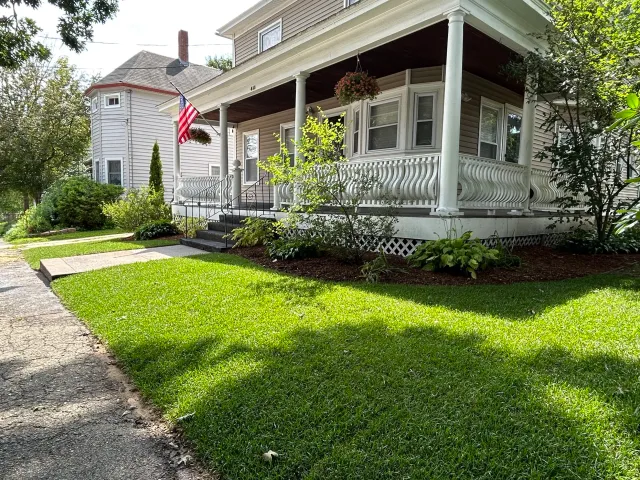 A suburban house with a porch and American flag, surrounded by a well-maintained lawn and foliage.
