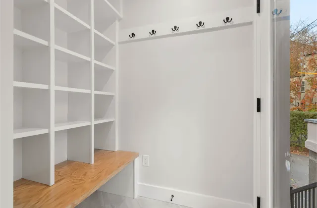 Expert design shines in this white mudroom featuring built-in cubbies, a wooden bench, wall hooks, and an open door to the outside.