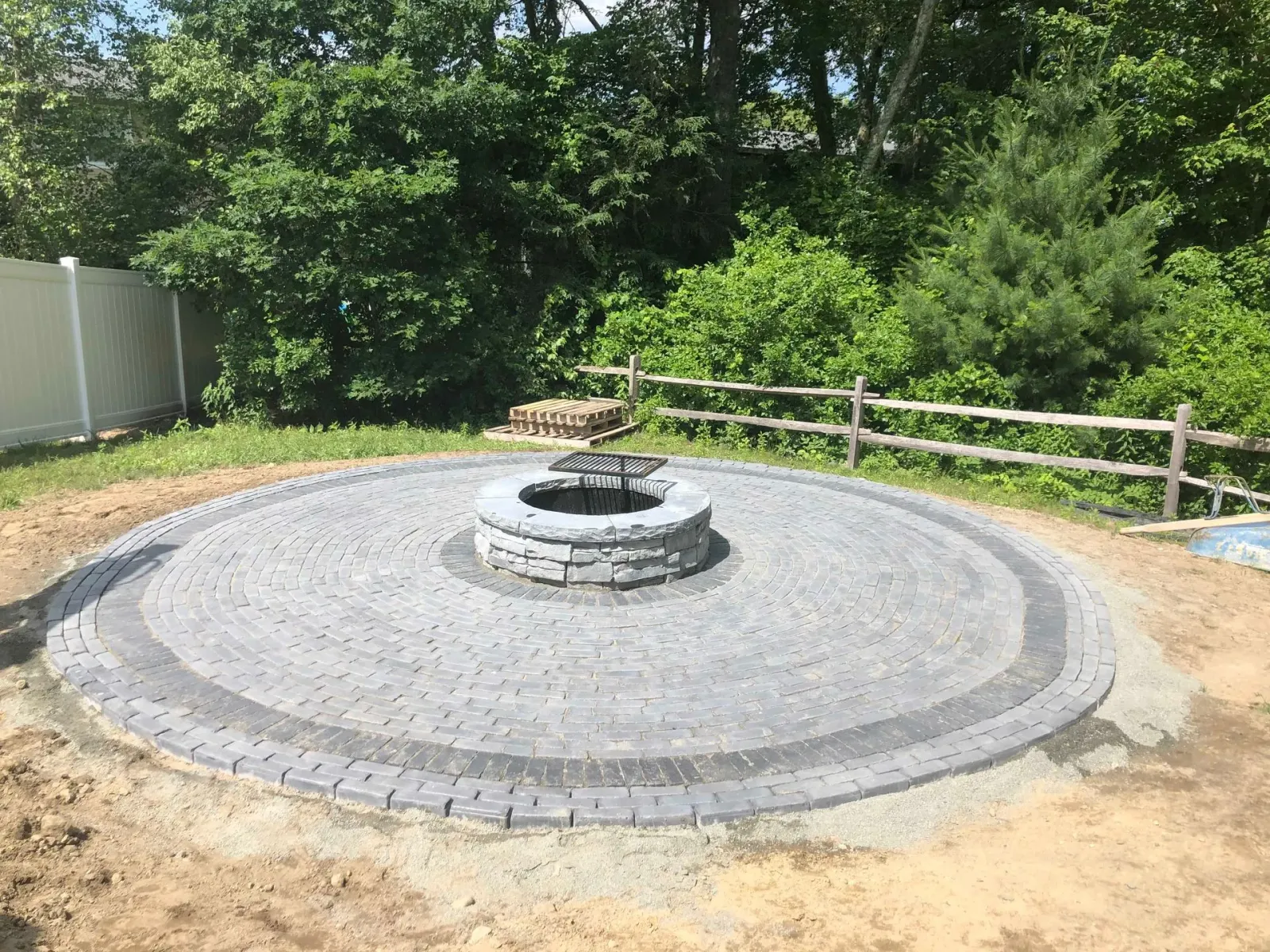A circular stone patio with a central fire pit sits in a grassy yard, surrounded by a split-rail fence and trees.