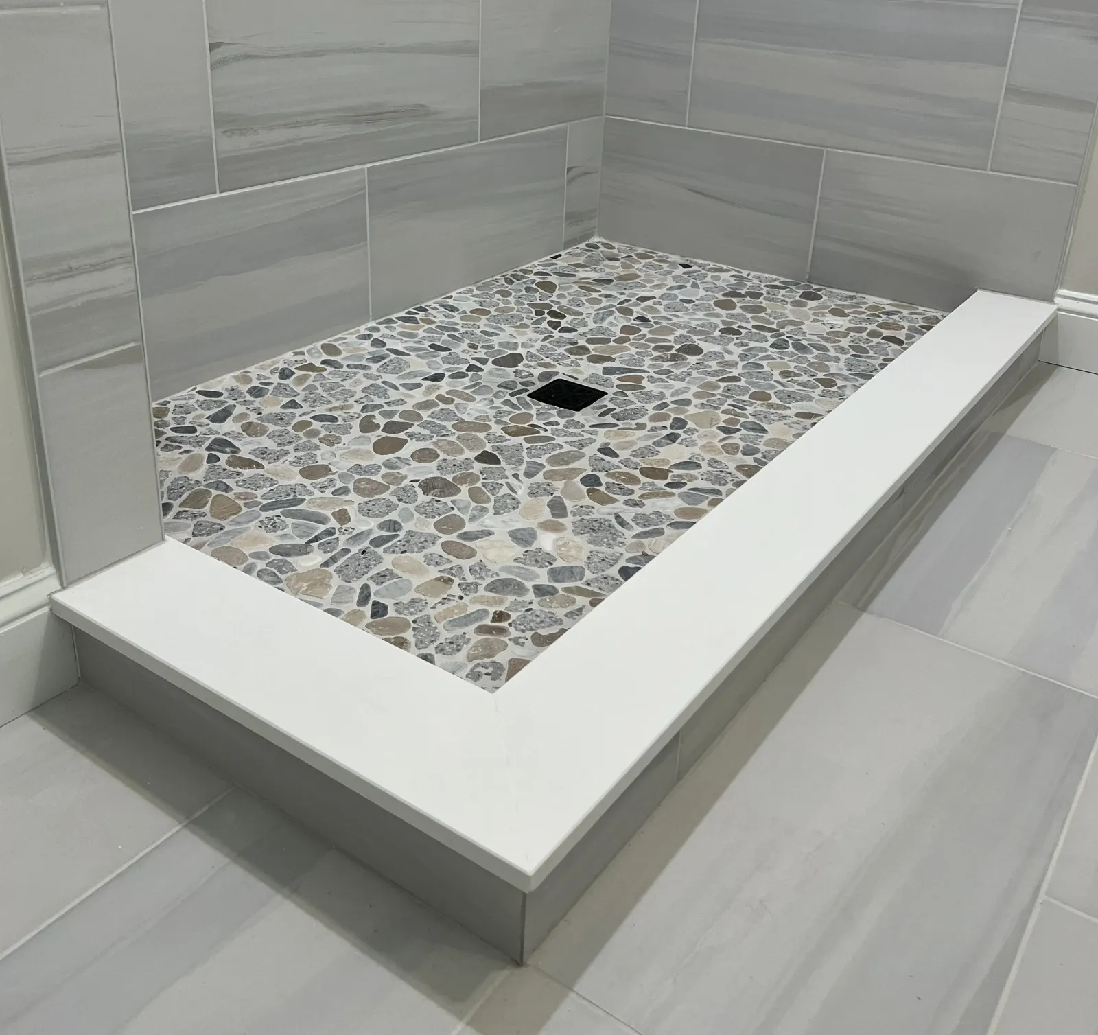 A modern shower base featuring a pebble stone design and a square drain, surrounded by sleek gray tiled walls and a white border.