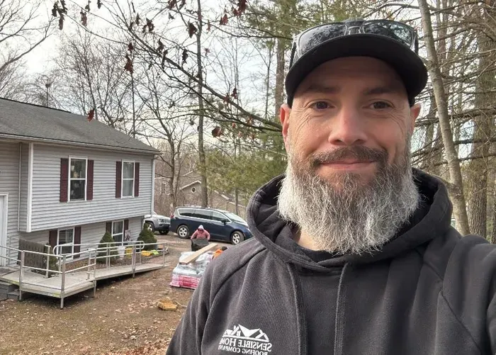 Man with grey beard and cap in front of a two-story house with a car and people in the background.