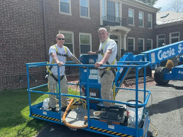 Two workers on a blue Genie lift outside a brick building, with tools and equipment on the platform.
