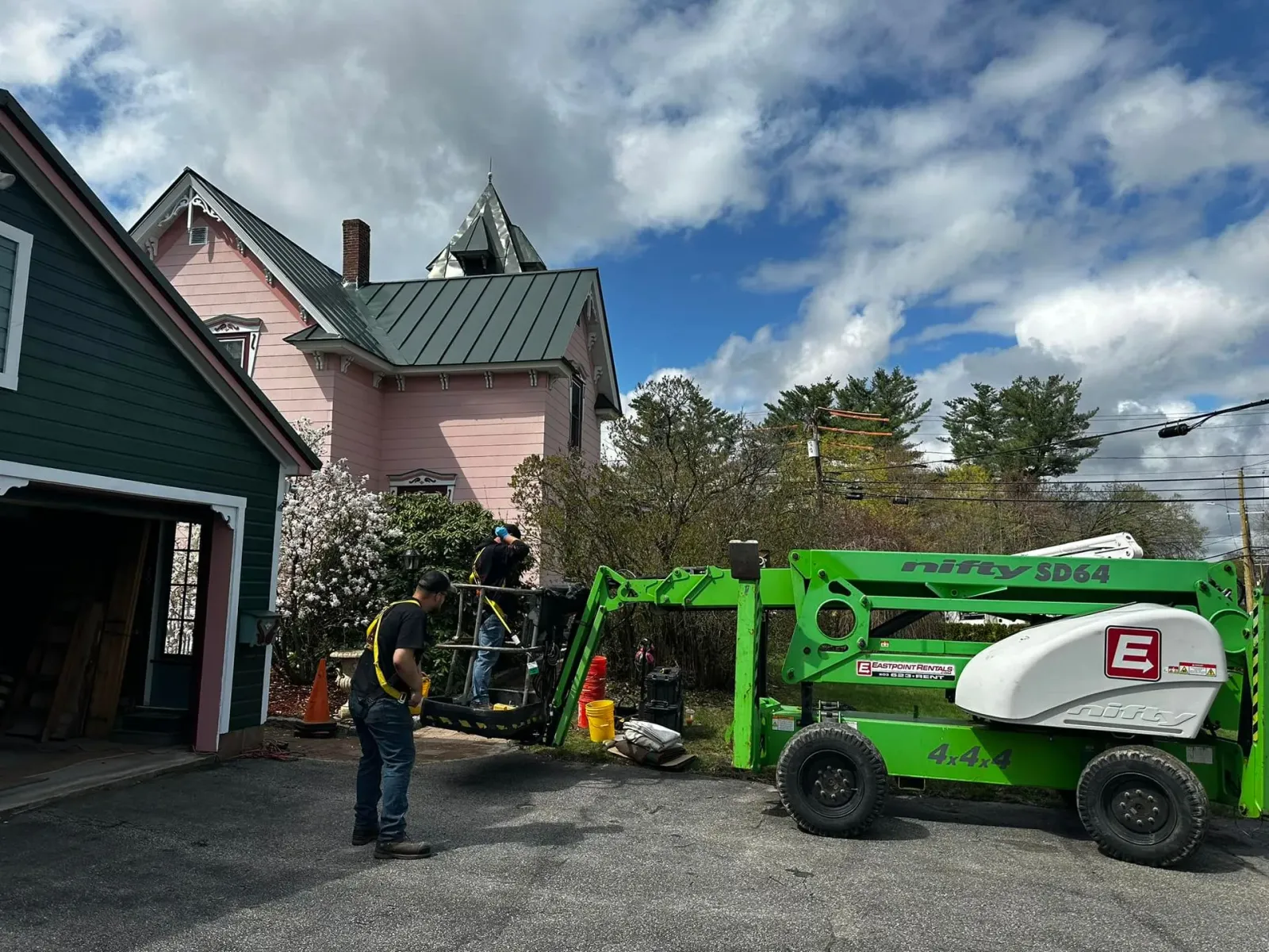 A worker in a harness near a green lift, pink house, and green garage on a cloudy day.