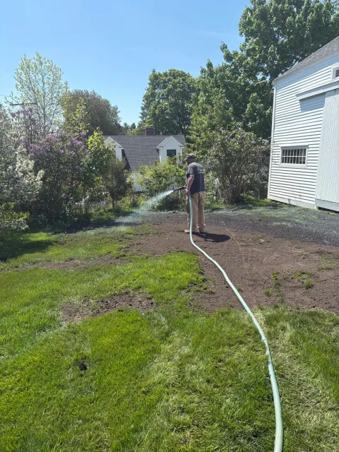 A man watering a garden with a garden hose next to a white house on a sunny day with clear blue sky and green trees.