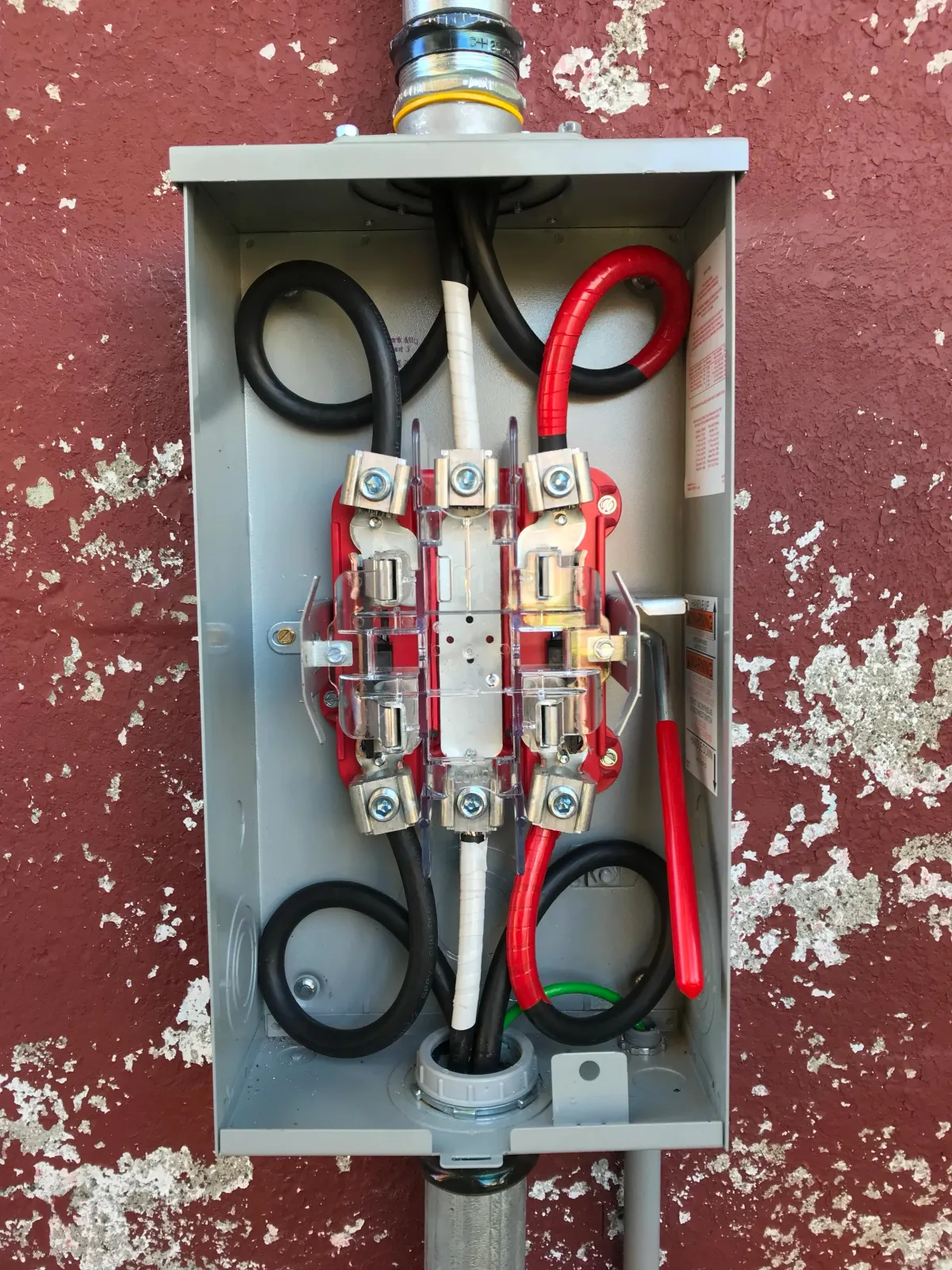 Open electrical panel with black and red cables connected to circuit breakers, mounted on a red wall.
