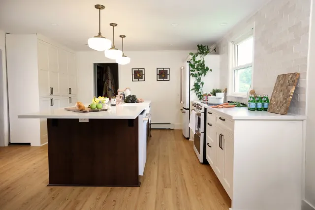 Bright modern kitchen with white cabinets, a dark wood island, and a white countertop. There are three pendant lights hanging above the island. A window overlooks the outside, with green plants near the window and a wooden cutting board with bottles of sparkling water on the countertop.