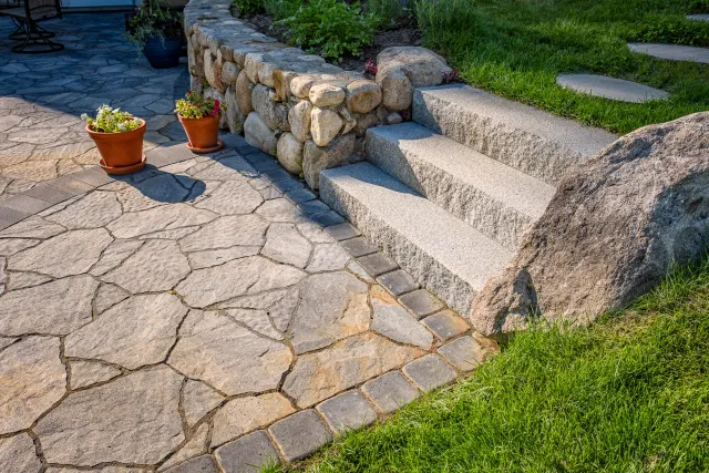 Patio with flagstone flooring, stone steps, rock wall, potted flowers, and grassy area.