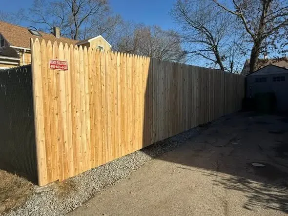 Wooden fence with pointed top along a paved area; sunny day.