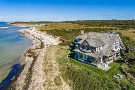 Aerial view of a modern coastal home on a grassy bluff overlooking a sandy beach and blue ocean waters