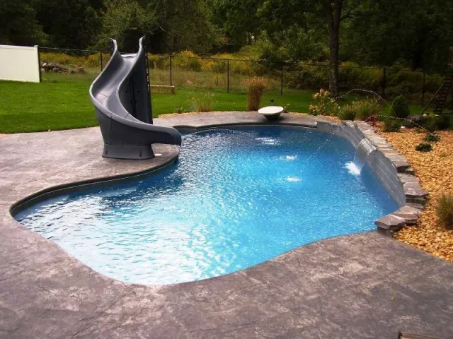 Backyard swimming pool with a water slide and a small ledge, surrounded by a concrete deck and landscaped yard with trees and plants.