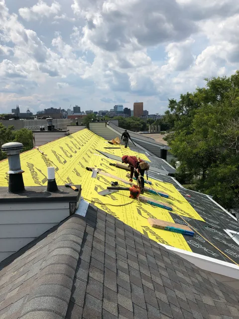 Storm King Roofing worker installing shingles over yellow TopShield underlayment on a Brookline, MA home with the Boston skyline in the background