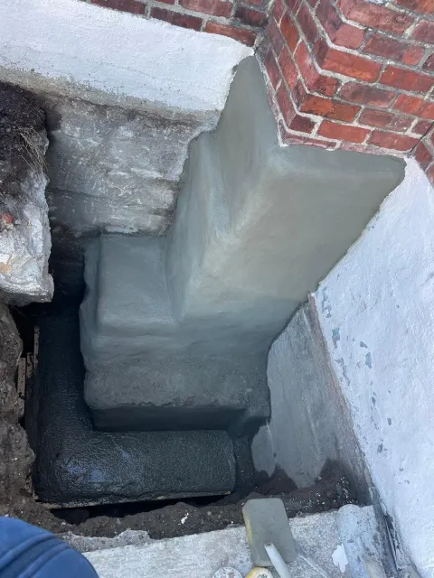 Newly cemented foundation repairs next to a brick chimney and white wall.