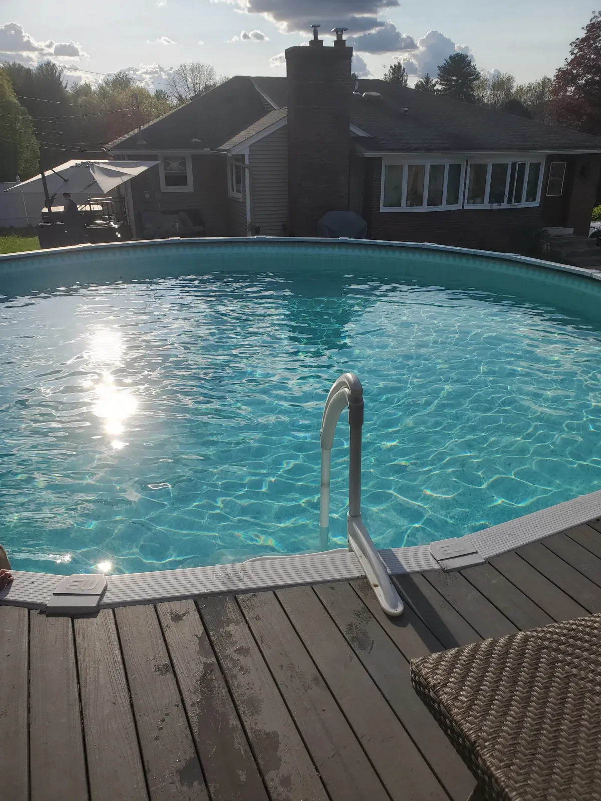 Backyard scene with a swimming pool, a wooden deck, wicker chair, an umbrella, house with large window and chimney, trees, and a partly cloudy sky.