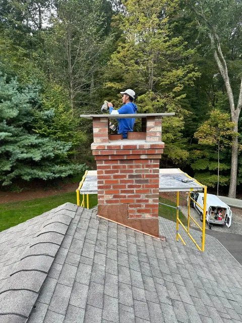 A worker in a blue shirt repairs a red brick chimney on a house roof next to yellow scaffolding and green trees.