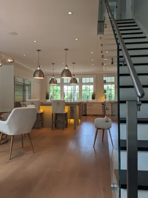 Modern kitchen with large window, three pendant lights above island, white cabinetry, hardwood flooring, glass staircase with metal railing.