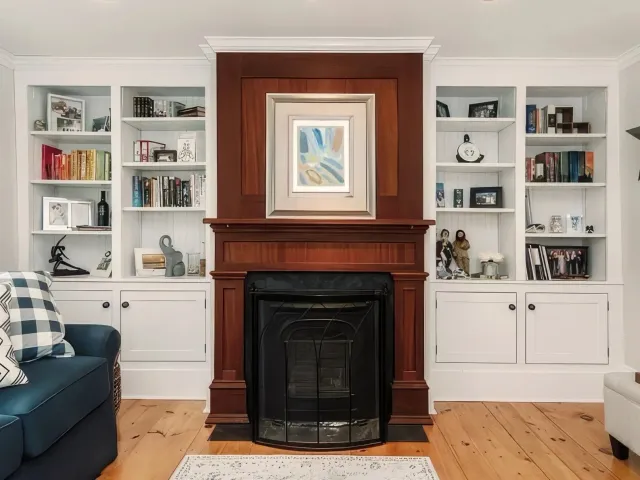 Living room with built-in white shelves on both sides of a fireplace, wood-paneled wall with a framed photo of a couple above the mantle, blue sofa with patterned pillows on the left, light-colored carpet on a wooden floor.