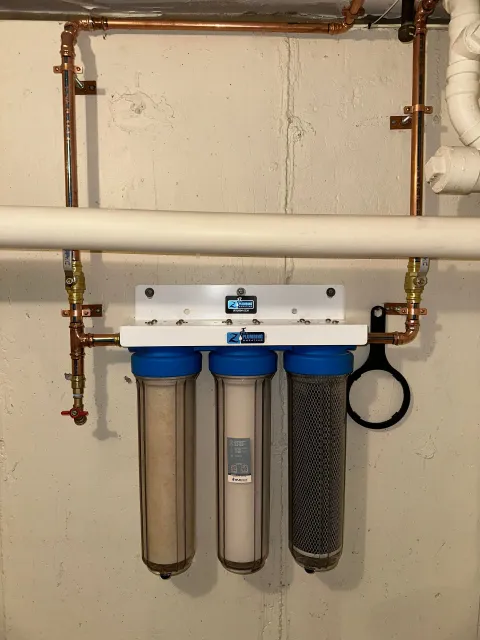 Water filtration system with three clear filter housings, copper piping, and white mounting plate on a concrete wall.