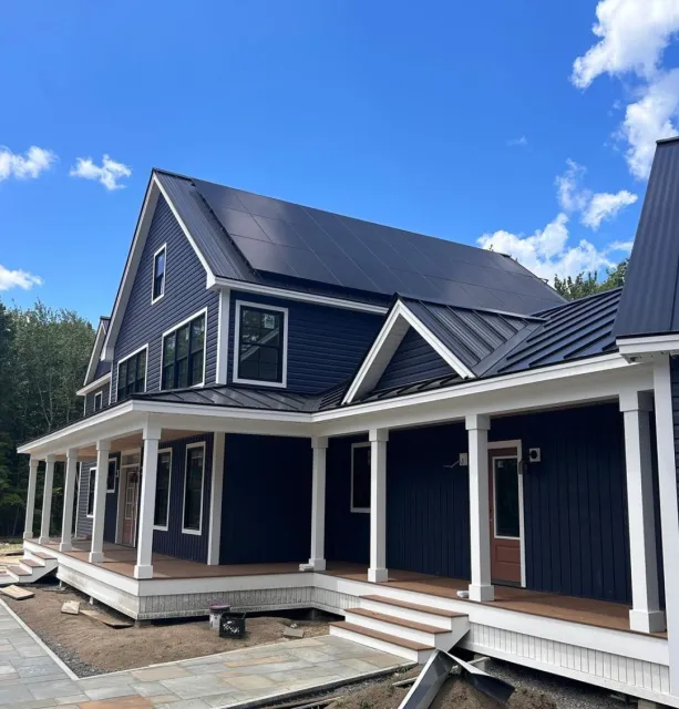 New two-story house with blue siding, white trim, a covered porch, and solar panels on the roof under a blue sky with clouds.