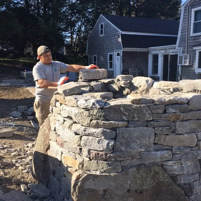 A person builds a stone wall near a small house, wearing work gloves and a cap, under clear skies in a rural setting.