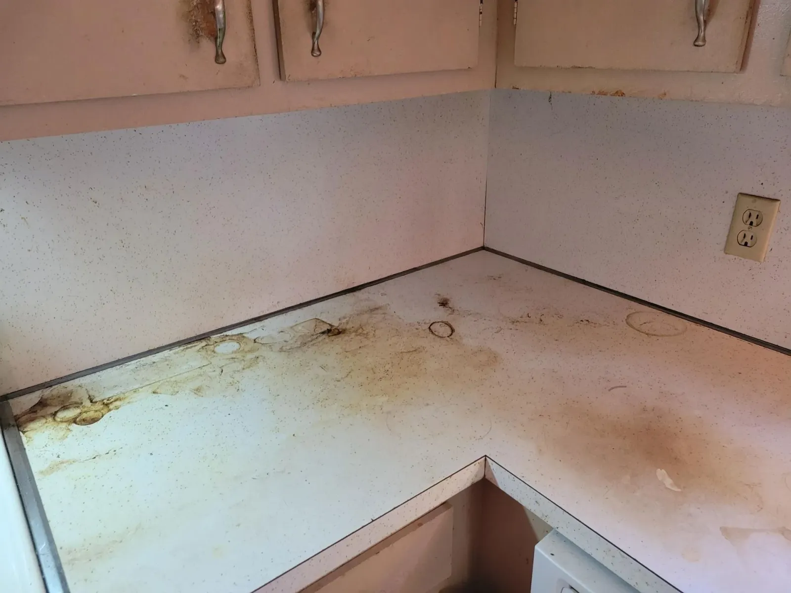 Dirty white kitchen countertop with brown stains, next to white cabinets and a wall outlet.