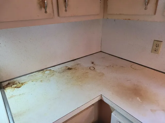Dirty white kitchen countertop with brown stains, next to white cabinets and a wall outlet.
