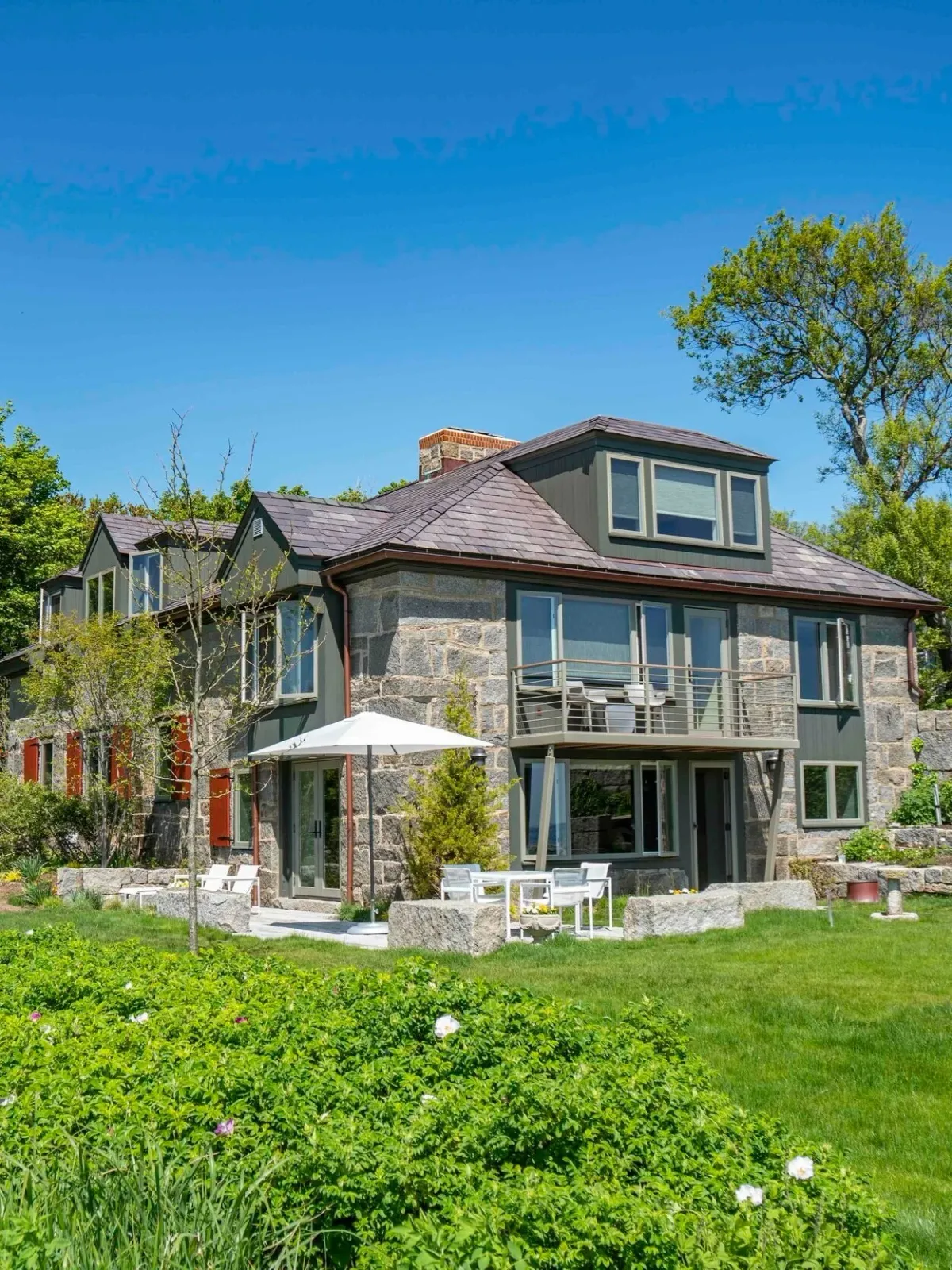 Historic granite home with a stone and green exterior, patio with white outdoor furniture, large umbrella, and lush greenery under a clear blue sky.