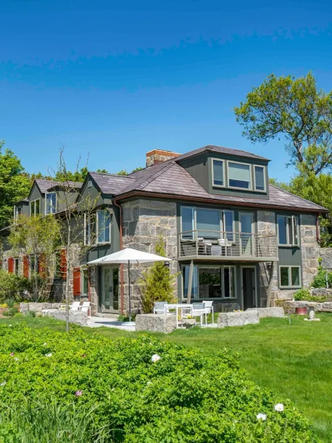 Historic granite home with a stone and green exterior, patio with white outdoor furniture, large umbrella, and lush greenery under a clear blue sky.