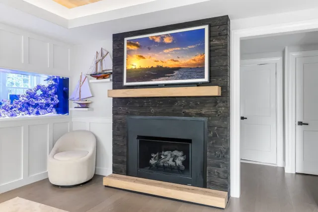 Living room with a wall-mounted TV displaying a sunset over the water, a black wooden fireplace with a light wooden mantel, white wainscoting, a beige round armchair. Fireplace and mantle installation by Watkins Works.