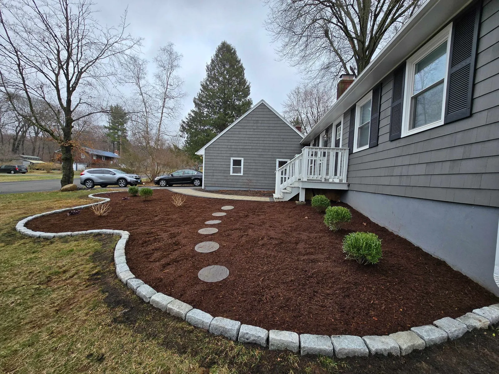 A gray house with new mulch, stone edging, and round stepping stones leading to a white deck on a cloudy day.