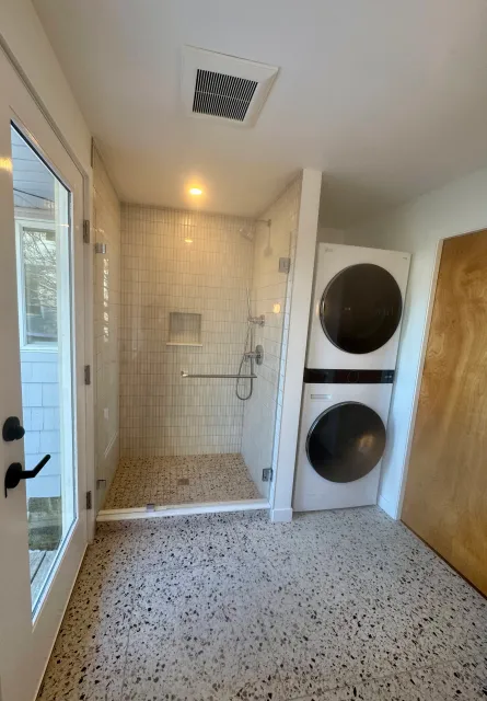 A small bathroom with a glass-enclosed shower, a washer and dryer stacked against the wall, a terracotta-tiled floor, and a door with a glass window leading outside.