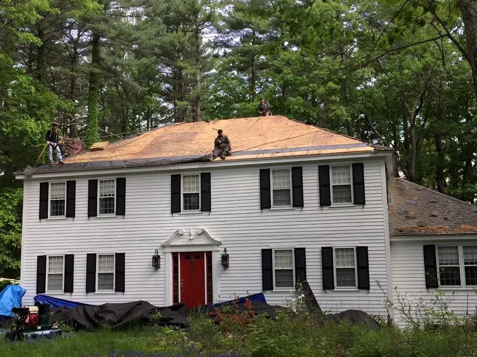 Roofers working on a white house with black shutters; red door. Trees in the background.