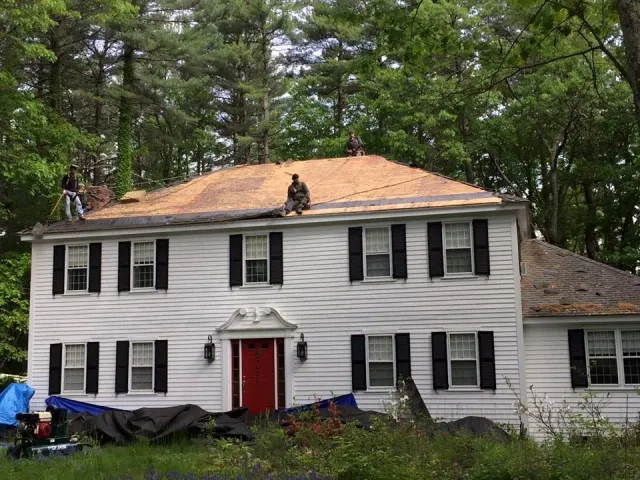 Roofers working on a white house with black shutters; red door. Trees in the background.