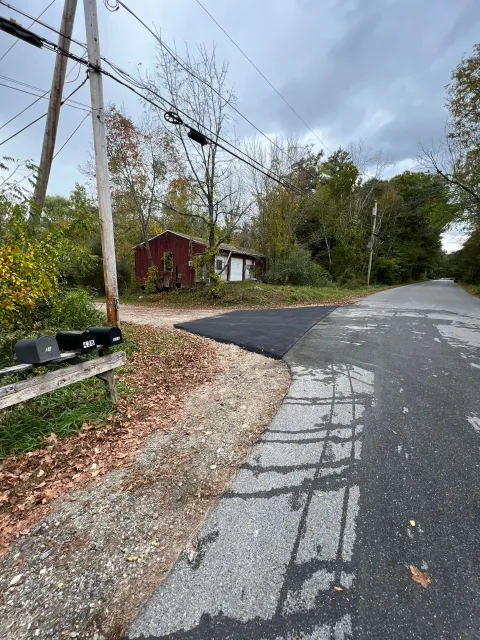Asphalt patch on a road by a gravel driveway; red shed visible amongst trees and power lines on a cloudy day.
