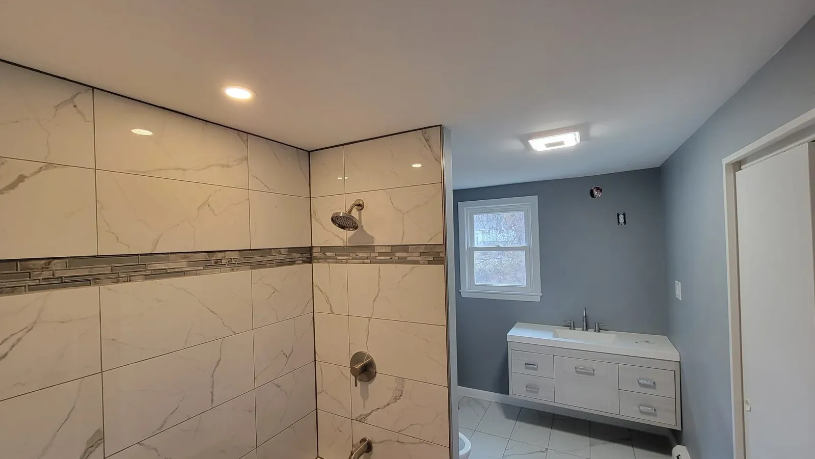 Modern bathroom with large tiled shower, recessed ceiling light, gray walls, white vanity with dual sinks, window, and tile flooring.