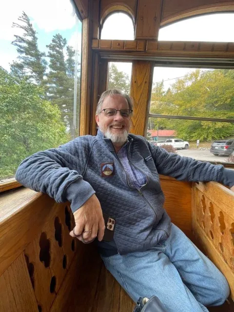 Smiling middle-aged man with glasses and a beard sitting inside a wooden structure with large windows, wearing a blue quilted jacket and jeans, with a scenic outdoor view of trees and parked cars.