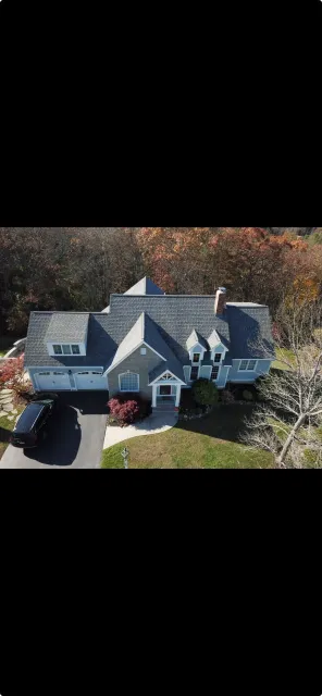 An aerial view of a large, multi-story house with a gray roof, white windows, a garage with a black car parked in the driveway, and surrounded by trees with fall foliage.