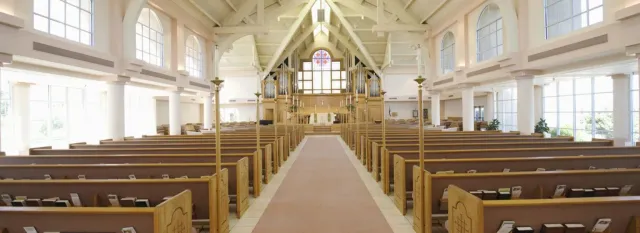 Interior view of a church with rows of wooden pews leading to the altar and a stained-glass window.