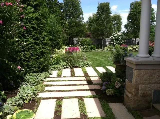 Stone stepping path through a lush garden with pink flowers and white columns.