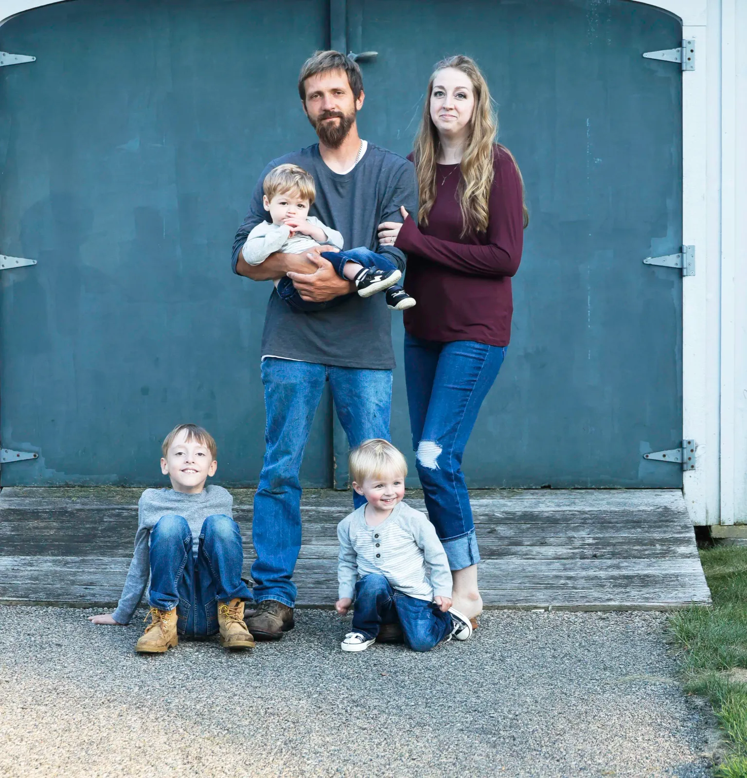 A family of five standing in front of a gray garage door. The father is holding a baby, and the mother is standing beside him. Two young children are sitting on the ground in front of them.