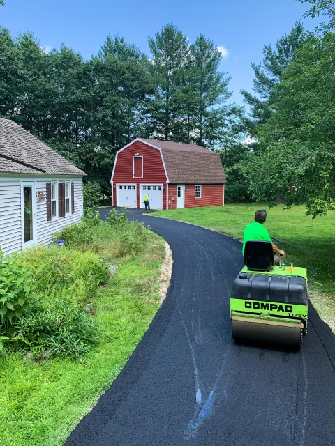 Person operating a lime green compaction machine on a freshly paved asphalt driveway, with another worker near a red barn in the background, during daytime with blue sky and trees.