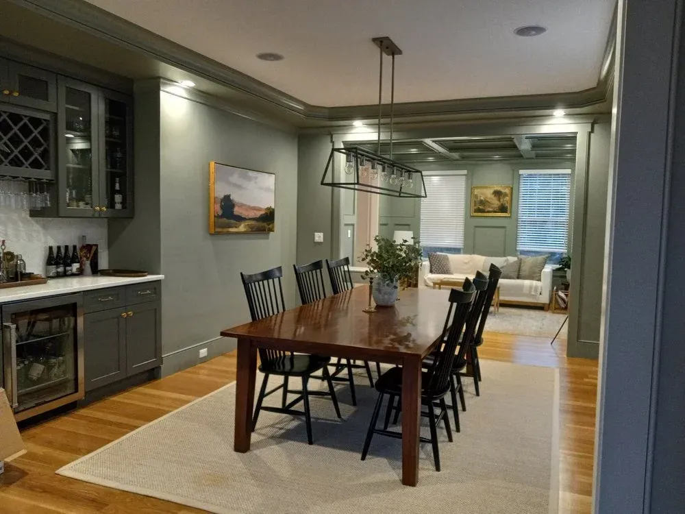 Dining area with a wooden table, eight black chairs, a floral centerpiece, and a wine bar with glassware and bottles, open to a living room with a white sofa and large windows.