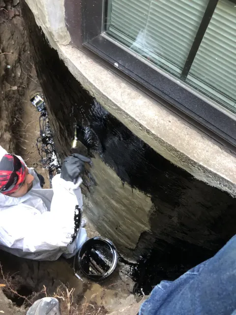 Worker applying waterproof coating to an exterior foundation wall during a basement waterproofing project