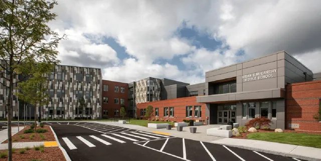 Modern school building with patterned facade, spacious parking area, and landscaped surroundings under a partly cloudy sky. No people visible.