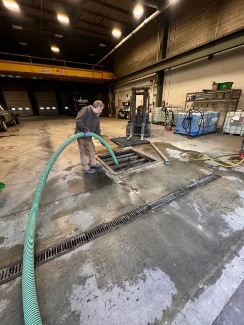 Man using a hose to clean a floor drain in a warehouse, near a forklift and equipment.