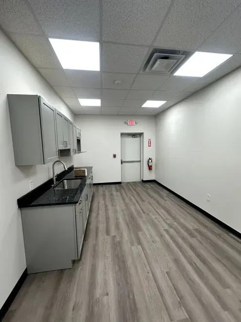 Empty break room with gray flooring, white walls, cabinets, countertop, and ceiling lights.