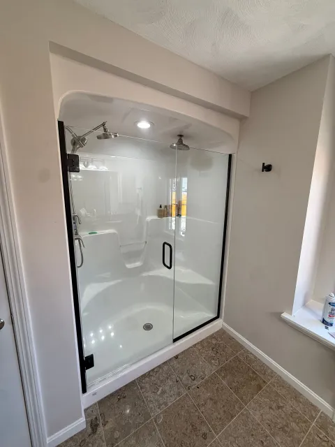 Glass walk-in shower with chrome fixtures in a beige bathroom corner, beside a window and carpeted floor.