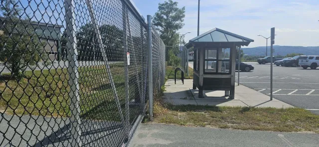 A small wooden guardhouse with glass windows is situated next to a parking lot, with a chain-link fence separating it from a grassy area, and mountains in the background on a sunny day.