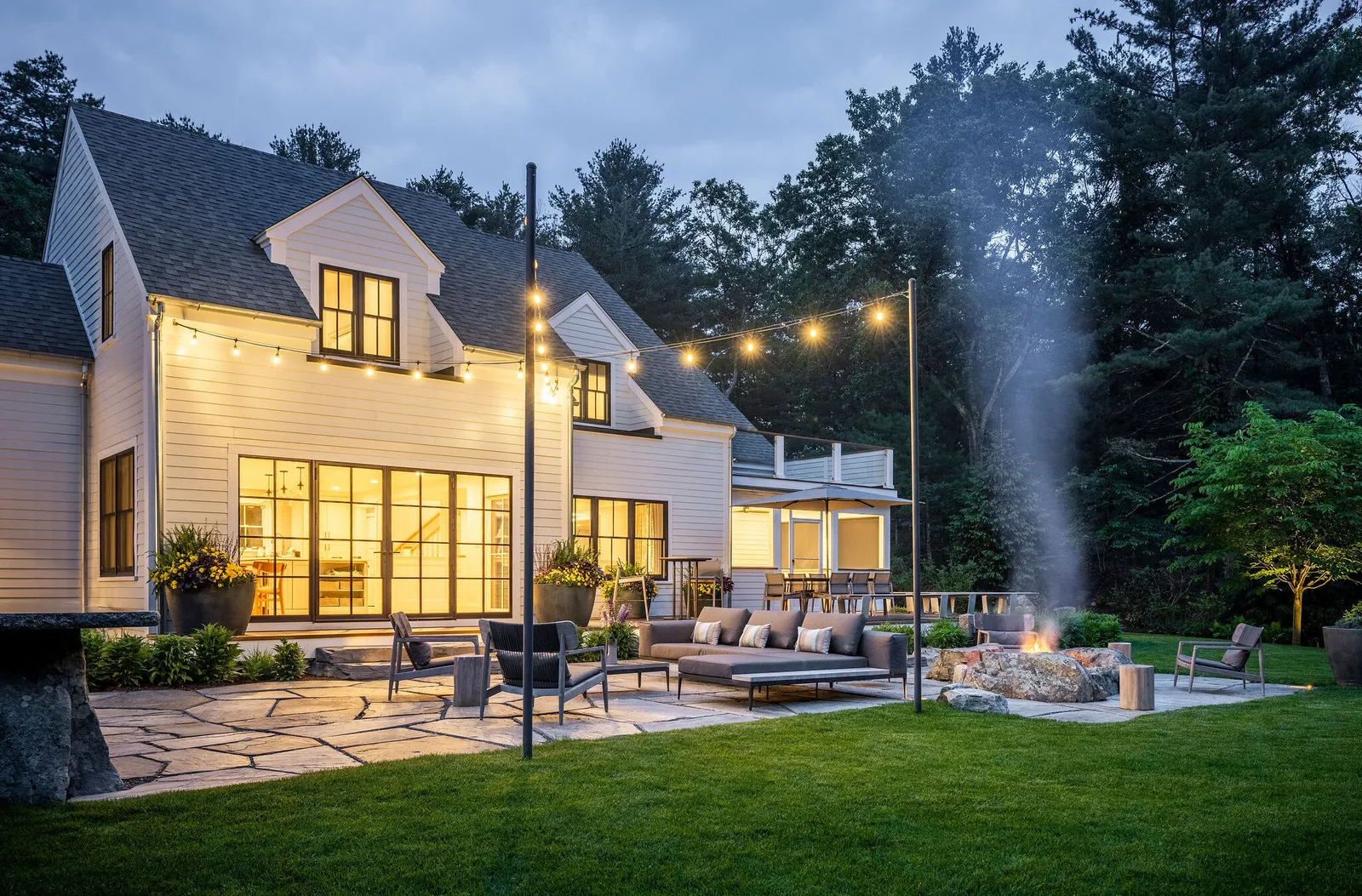 This image shows a modern two-story home at dusk, with outdoor lighting, a fire pit, lounge furniture, and a lush lawn surrounded by trees.